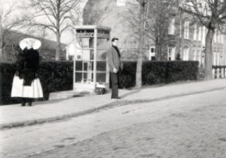 Een man staat buiten bij een telefooncel en een vrouw in Zuid-Bevelandse strekdracht staat twee meter verderop