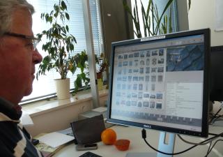 Rinus kijkt naar het beeldscherm van zijn computer, waarop pictogrammen van foto's te zien zijn