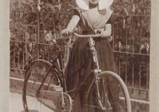 Vrouw in streekdracht houdt haar fiets vast aan het stuur. Er zit een gehaakte jasbeschermer op het achterwiel