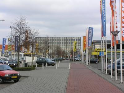 Marconistraat met in de verte het Stadskantoor en links en rechts aan beide zijden van een fietspad staan auto's geparkeerd, vlaggen aan lange masten staan erbij om de namen van winkels aan te geven