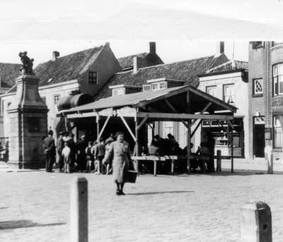 Mensen verzamelen zich op een marktplein met een kraampje, één vrouw loopt net weg