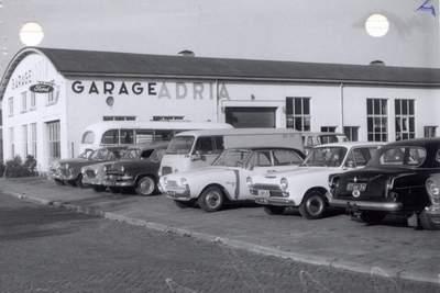 Loods met de tekst "Garage Adria" groot geschreven. Ernaast staat een parkeerplaats vol auto's