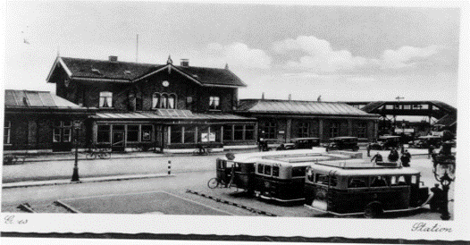 Stationsgebouw met parkeerterrein ervoor. Er staan drie bussen geparkeerd, en vlak voor het station zelf ook nog een rij met auto's