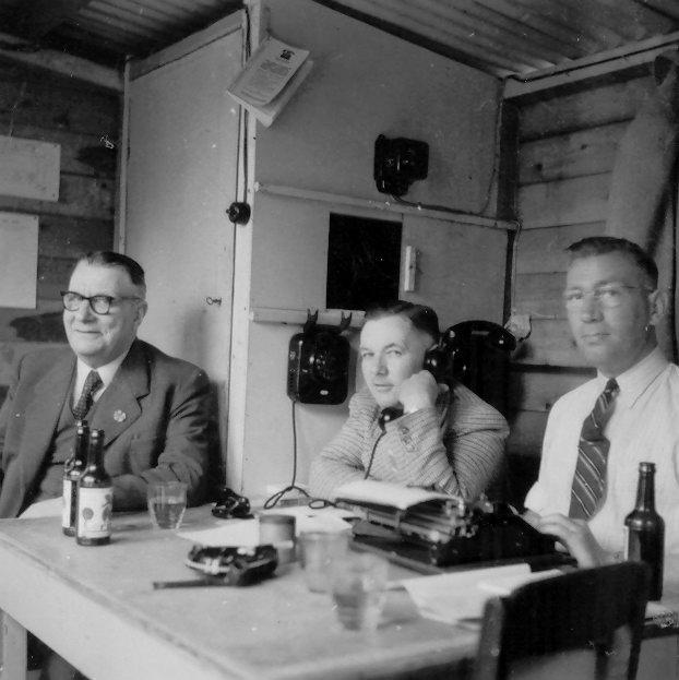 3 mannen zitten een een tafel, één is aan het telefoneren