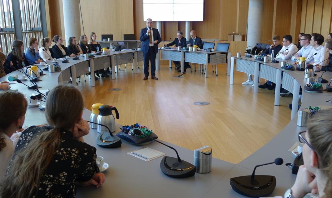 Raadszaal met wethouder André van der Reest. Leerlingen zitten aan de omringende tafels