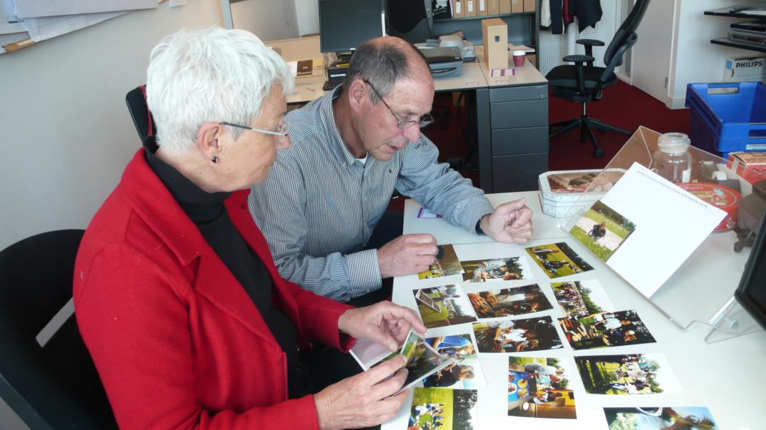 Adi en Toos zitten aan een tafel waarop veel foto's liggen