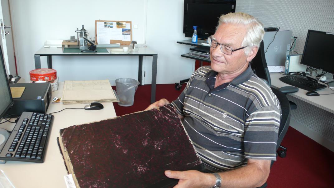 Huub Bellaart houdt een bevolkingsregister vast terwijl hij achter een bureau zit