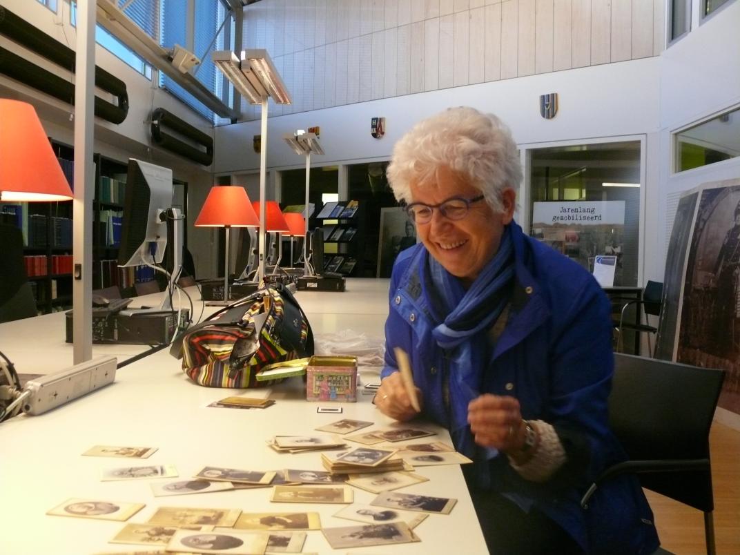 Nel Minnee in de studiezaal. Voor haar liggen op tafel allemaal portretfoto's uitgestald. 