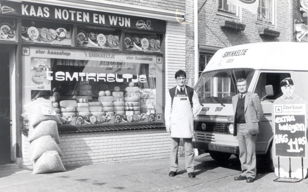 Winkel met reclamebord "kaas noten en wijn". 2 mannen staan buiten de zaak bij een busje