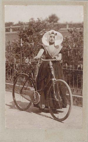 Vrouw in streekdracht houdt haar fiets vast aan het stuur. Er zit een gehaakte jasbeschermer op het achterwiel