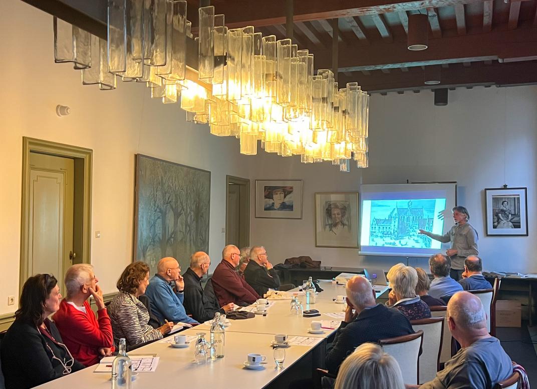 Frank geeft een lezing in een zaal in het stadhuis. Voor hem staat een lange tafel waar mensen aan zitten